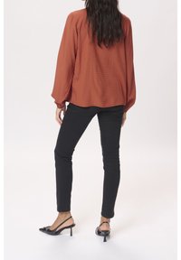 Rustfarvet bluse med teksturerede prikker, løs pasform og lange ærmer, parret med sorte skinny jeans og sorte hæle.
