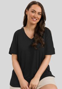 Donna sorridente con lunghi capelli castani ondulati indossa una maglietta nera a V e pantaloni beige, seduta contro uno sfondo grigio semplice.