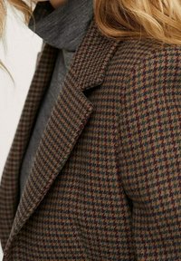 Blazer de patrón houndstooth en marrón y verde con cuello de muesca, que presenta una tela texturizada y un ajuste entallado. Suéter de cuello alto gris debajo.