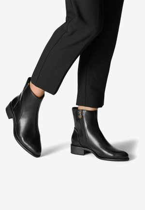 Personne portant des bottines noires avec des fermetures éclair latérales et un pantalon noir coupé au-dessus de la cheville, debout sur une surface claire.