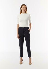 Weißes kurzärmeliges Oberteil kombiniert mit dunkel gestreiften Hosen und beigen spitzen Pumps. Das Outfit zeichnet sich durch eine taillierte Form und eine glatte Textur aus.