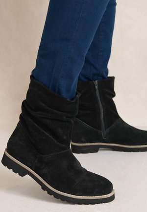 Bottes mi-mollet en daim noir avec fermeture éclair latérale portées avec un jean skinny bleu foncé sur fond neutre.