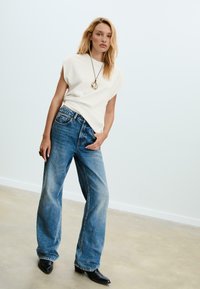 Haut léger beige ample à manches courtes, associé à un jean en denim bleu à pattes larges et des bottes noires à bout pointu, se tenant devant un fond clair.