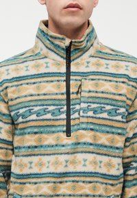 Άνδρας που φοράει φούτερ από φλις με ψηλό γιακά, σε γεωμετρικά σχέδια σε τόνους τιρκουάζ, μπεζ και κρεμ, με μαύρο φερμουάρ τεταρτημόριου μήκους και φερμουάρ σε τσέπη στο στήθος.