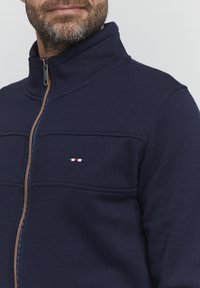 Giacca a zip blu navy con colletto alto, caratterizzata da un logo ricamato in rosso, bianco e blu, e una cerniera a contrasto. Tessuto morbido.