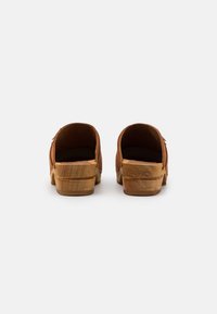 Zoccoli in suede marrone con base in legno, caratterizzati da una forma arrotondata e un morbido rivestimento interno, progettati per comfort e resistenza.