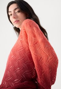Pull en tricot avec un dégradé de rouges et corails, arborant un motif ondulé et des manches larges et amples. Texture douce et légère.