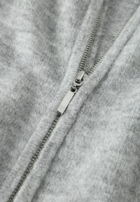 Vaaleanharmaa fleece-takki, jossa metallinen vetoketju ja vetoketjun vedin, pehmeä, teksturoitu pinta ja tiiviisti ommellut saumat.