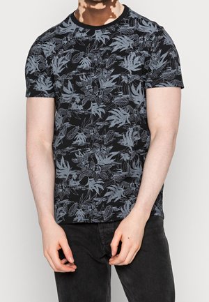 Mand iført sort t-shirt med kortærmer med gråt blomsterprint og sorte bukser, stående med hænderne løst foldet foran.