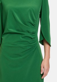 Grünes Kleid aus glattem Stoff mit seitlicher Raffung, weiten Ärmeln und einer eleganten Silhouette. Die Textur wirkt weich und fließend.