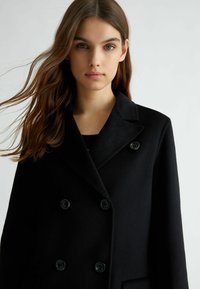 Cappotto nero doppiopetto realizzato in tessuto strutturato, con colletto a scialle e bottoni neri. Vestibilità sartoriale con tasche laterali.