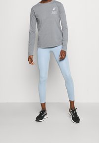 Graues Langarmshirt mit weißem Schriftzug, hellblaue Leggings und schwarze Sportschuhe mit weißen Sohlen und bunten Akzenten.