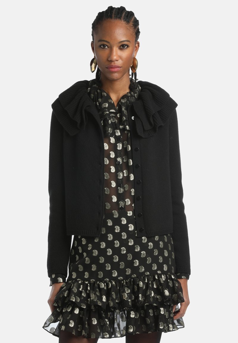 Schwarze Strickjacke mit Rüschensaum und Knopfverschluss. Tragen über einem transparenten schwarzen Kleid, das mit goldenen kreisförmigen Mustern verziert ist.