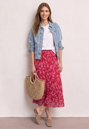 Lächelnde Frau mit langen Haaren trägt eine helle Jeansjacke, ein weißes Hemd, einen roten gemusterten Midirock, beige Slipper und hält eine geflochtene Handtasche.