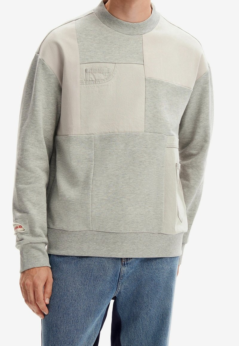 Homme portant un sweat-shirt patchwork gris avec une petite poche poitrine et un jean bleu clair, debout contre un fond blanc.