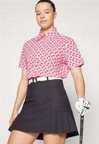 Short-sleeve polo shirt in pink with white geometric pattern, paired with a black pleated skirt and a white belt, holding a golf club.