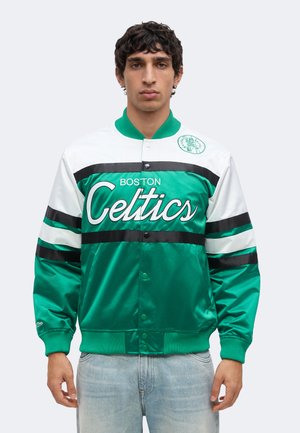 Άνδρας που φοράει ένα σατέν μπουφάν Boston Celtics σε πράσινο και άσπρο με μαύρες ρίγες και ανοιχτό μπλε τζιν, στέκεται μπροστά σε απλό φόντο.