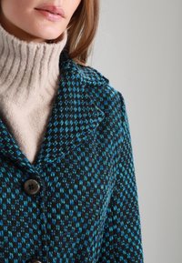 Cappotto in tessuto con un motivo a quadri nero e blu, dotato di colletto, chiusura a bottoni e un dolcevita beige testurizzato sotto.