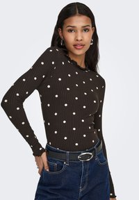 Brun ribstrikket langærmet top med hvide prikker, med en flæsekant ved halsudskæringen. Kombineret med højttaljede blå jeans og et sort bælte.