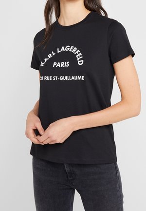 Kobieta w czarnej koszulce Karl Lagerfeld Paris z białym napisem i ciemnych dżinsach, ręce złożone z przodu.