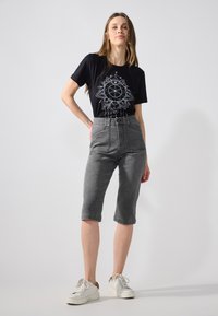 Fekete póló fehér geometrikus grafikával, magas derekú szürke farmer capri nadrággal párosítva. A modellen fehér sportcipő van.