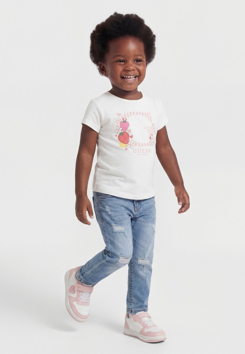 Bambin souriant marchant, portant un t-shirt blanc avec un imprimé de fruits, un jean bleu déchiré, et des baskets roses et blanches.