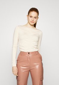 Blusa de mangas longas, bege claro, com detalhes franzidos à frente, combinada com calças cargo de cintura alta, brilhantes e cor-de-rosa, com bolsos.