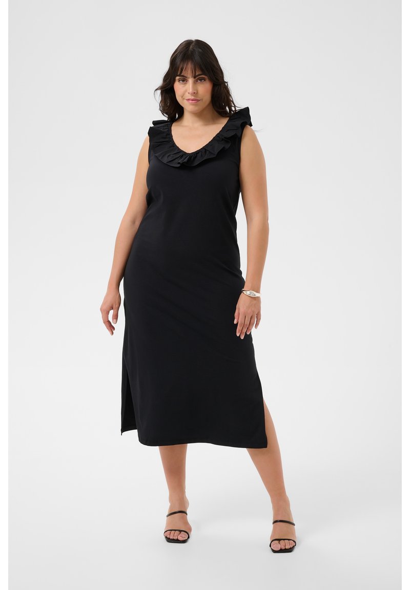 Femme portant une robe noire sans manches avec un col à volants et une fente latérale, debout avec un pied en avant sur un fond uni.