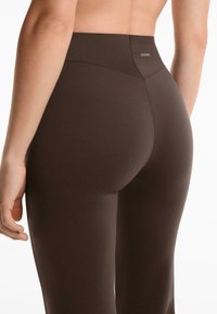 Rückenansicht einer Person in hoch taillierten, dunkelbraunen, enganliegenden Leggings mit dem Oysho-Logo in der Mitte des Bundes.