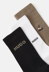 Drei Paar gerippte Socken in Schwarz, Weiß und Braun, jeweils mit "HUGO"-Markenname; das braune Paar hat oberhalb des Logos einen kleinen gestickten Vogel.