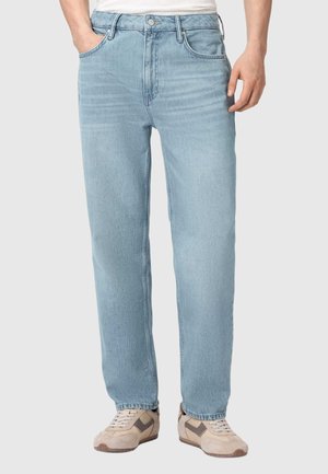 Man draagt lichtblauwe rechte jeans, één hand in de zak, gecombineerd met beige en bruine casual sneakers op een witte achtergrond.