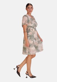 Vestido floral con un patrón verde y rosa, mangas cortas, cintura fruncida y una falda fluida. Combinado con zapatos de tacón alto negros y brillantes.