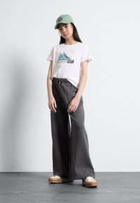 T-shirt blanche avec motif de baskets turquoise, pantalon large gris avec motif texturé et casquette vert clair. Baskets beiges avec semelles marron.