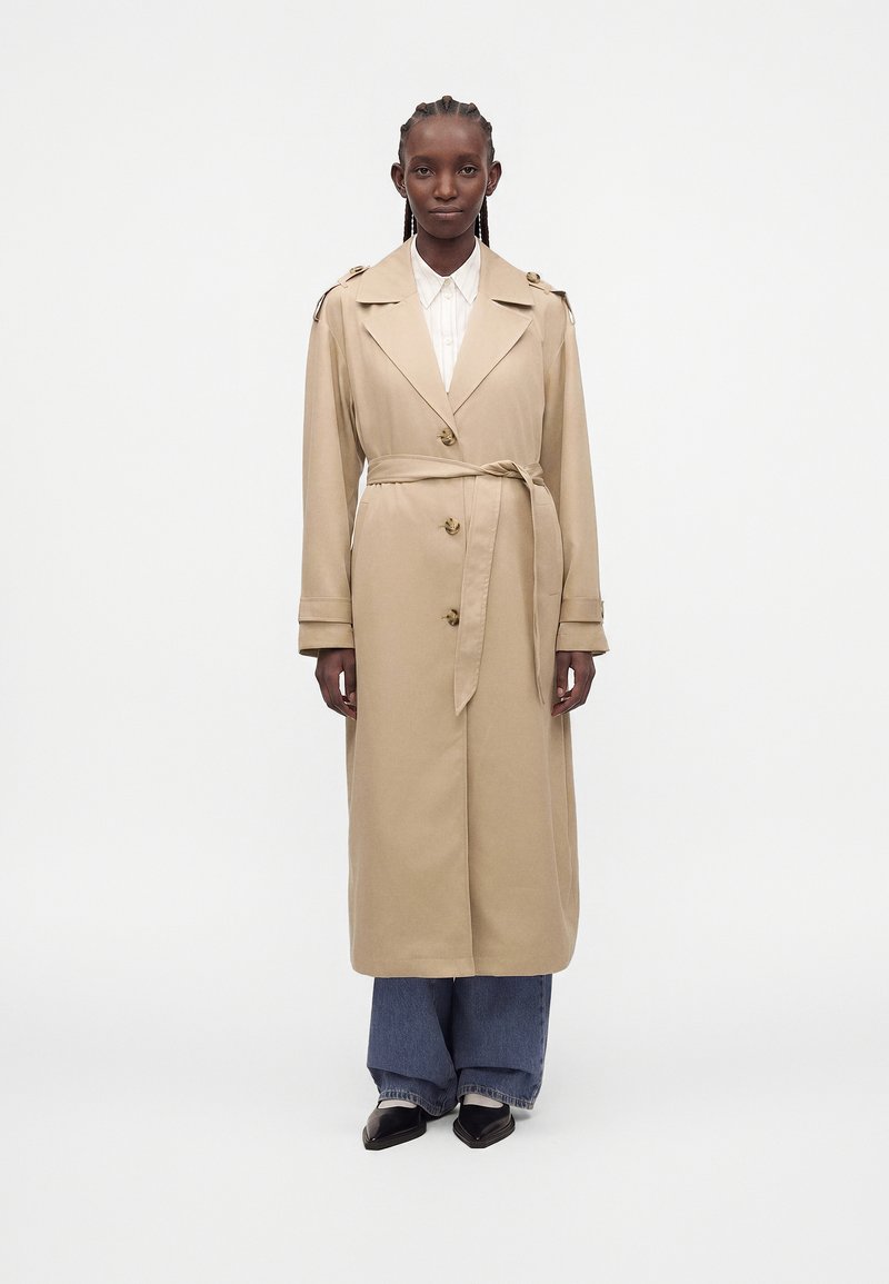 Personne grande portant un trench-coat beige ceinturé par-dessus une chemise blanche, un jean large bleu et des chaussures noires à bout pointu, debout devant un fond blanc uni.