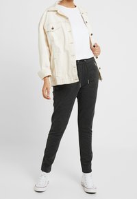 Krämfärgad denimjacka med knäppning och två bröstfickor, bärs över en vit t-shirt, i kombination med mörkgrå joggers och vita sneakers.