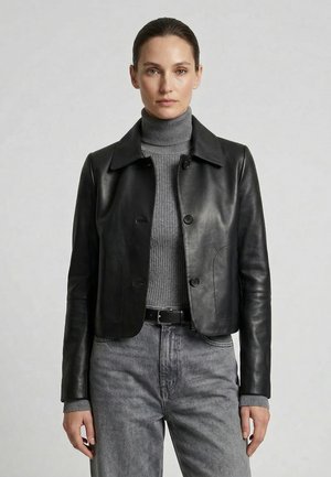 Vrouw in een zwarte leren jas met knopen over een grijze geribde coltrui, grijze jeans met riem, staand met een neutrale uitdrukking.