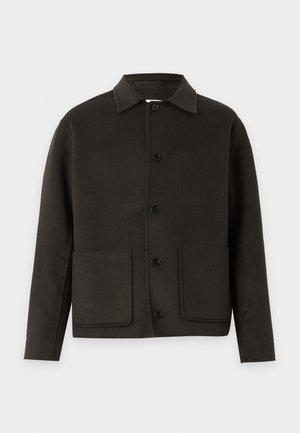Homme portant une veste noire boutonnée, une chemise blanche, un pantalon noir et des chaussures noires, debout devant un fond blanc uni.