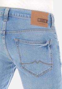 Vaqueros de denim azul claro con una etiqueta de cuero marrón etiquetada "MUSTANG True Denim." Presentan costuras en contraste y un solo bolsillo trasero con un diseño curvado.