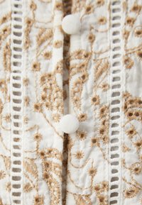 Valkoinen kangas, jossa on vaaleanruskeita brodeerattuja kukkakuvioita, ja siinä on kaksi beigeä nappia sekä pystysuoraa pitsiyksityiskohtaa reunoilla.