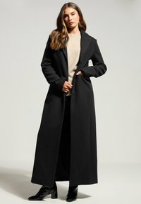 Cappotto di lana nero con un design lungo e dritto e colletto a patta. Presenta due tasche frontali ed è indossato sopra un maglione beige.