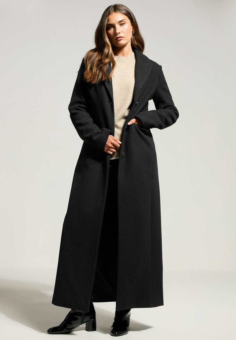 Cappotto di lana nero con un design lungo e dritto e colletto a patta. Presenta due tasche frontali ed è indossato sopra un maglione beige.