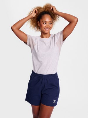 Uśmiechnięta kobieta w jasnoszarej koszulce i granatowych szortach poprawia swoje kręcone włosy na jednolitym białym tle.