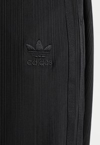 Pantalones deportivos negros con textura acanalada, que cuentan con un logo de Adidas negro bordado y franjas laterales negras como detalle.