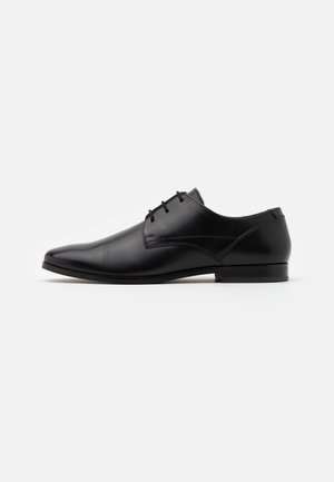 Chaussure habillée en cuir noir avec un bout pointu, un laçage, et de subtils accents texturés sur le dessus. Talon plat et profil minimaliste.