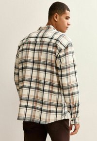 Hombre vestido con una camisa de cuadros en tonos beige, negro y marrón de manga larga, de perfil frente a una pared clara y lisa.