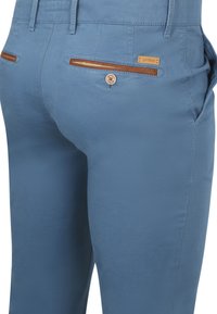 Helleblaue Hosen aus glattem Stoff, mit einem Knopfverschluss, Lederakzenten an den Taschen und einem rechteckigen Markenlabel.
