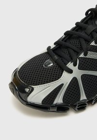 Baskets en mesh noir avec des accents en noir brillant et en argent, une tige texturée et des éléments de design distincts avec un capuchon de chaussure rigide.