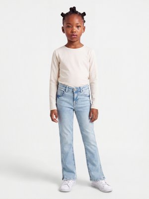 Jeune fille avec une coiffure en nœuds bantu, portant un t-shirt crème à manches longues, un jean bleu clair et des baskets blanches, debout sur fond blanc.