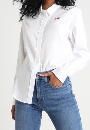 Mujer con camisa blanca de botones con el logo de Levi's y jeans azules, una mano en el bolsillo, de pie contra un fondo blanco.