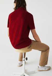 Polo bordeaux à manches courtes, coupe décontractée, associé à un pantalon kaki. Chaise blanche avec une assise texturée et chaussures grises.
